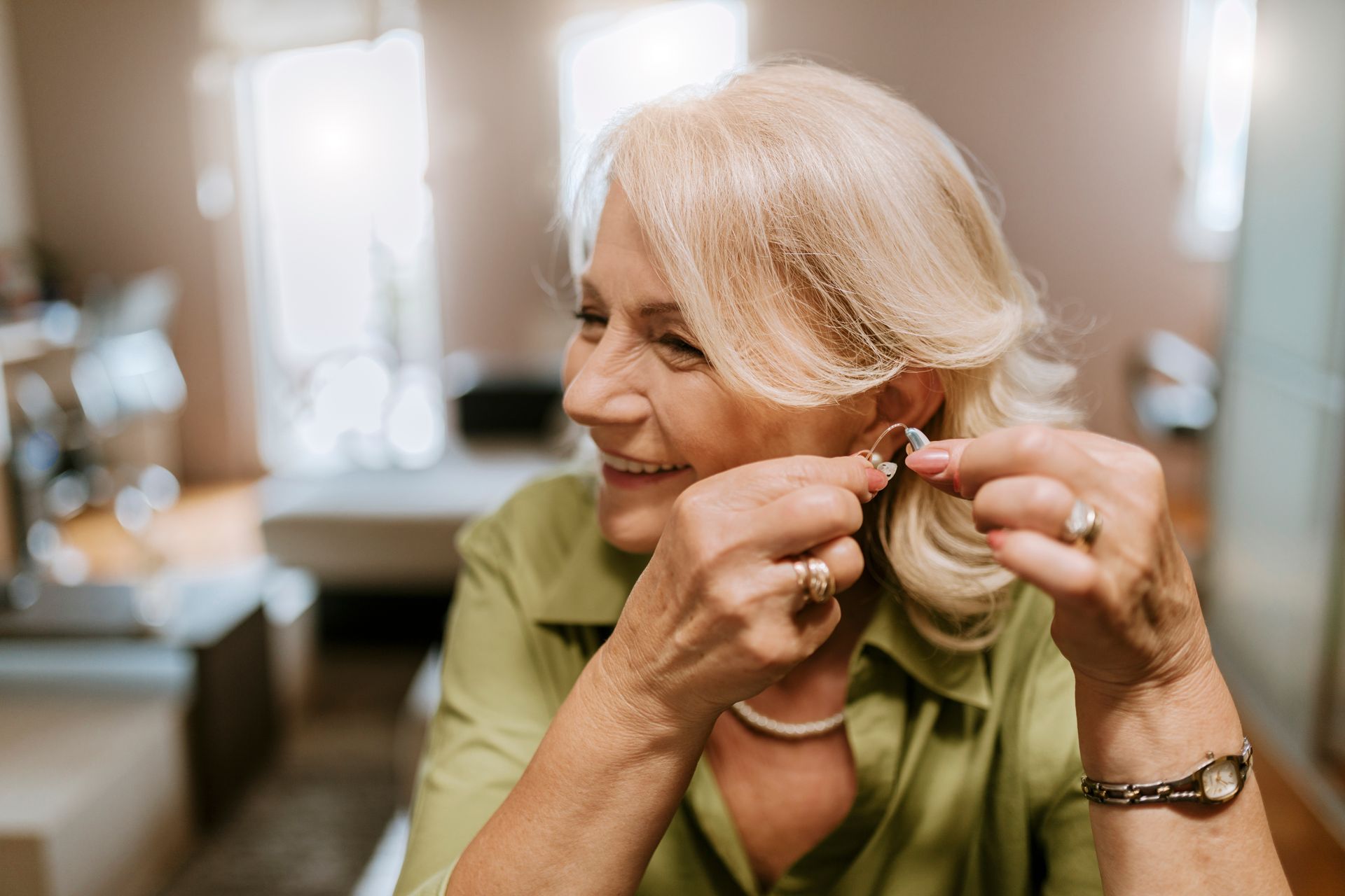 A smiling senior woman is fitting a hearing aid into her ear.