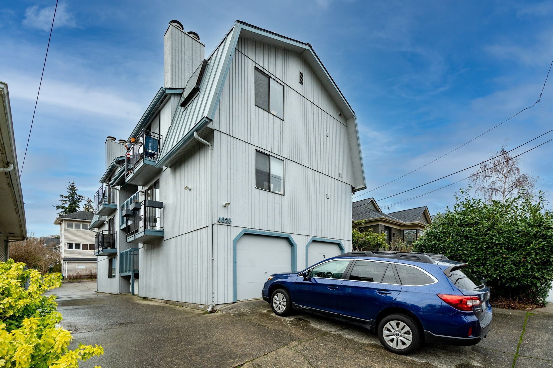 White multi-story house with balconies beside a blue SUV on a driveway under a clear sky