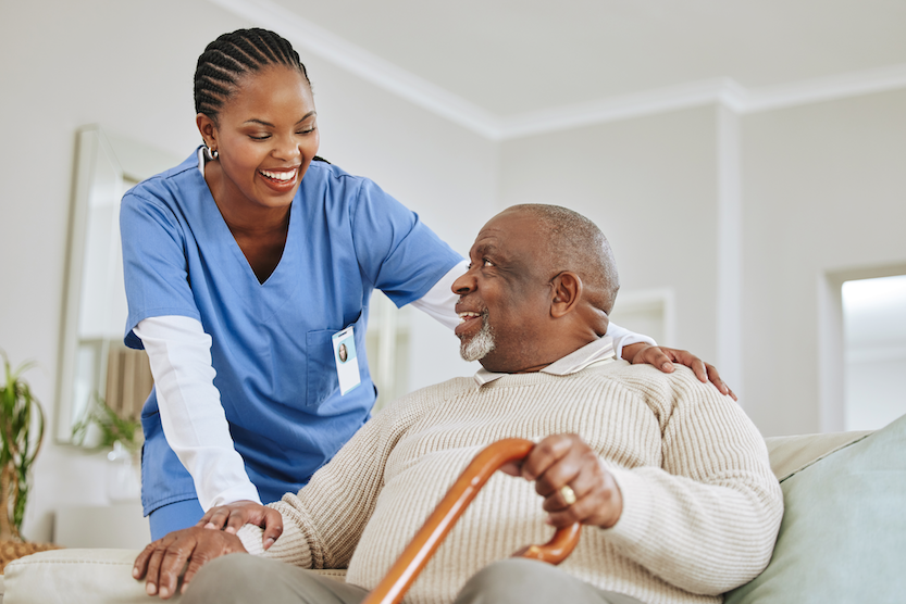 Caregiver in blue scrubs smiling and assisting an elderly man holding a cane while seated on a couch, featured in a blog article about home modifications for people with dementia.