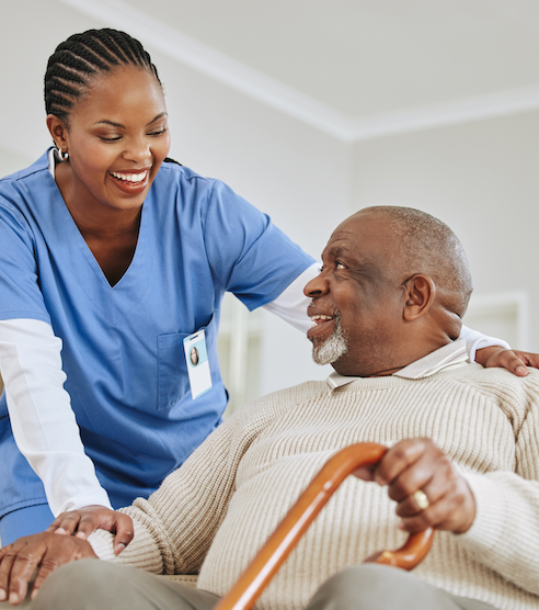 Caregiver in blue scrubs smiling and assisting an elderly man holding a cane while seated