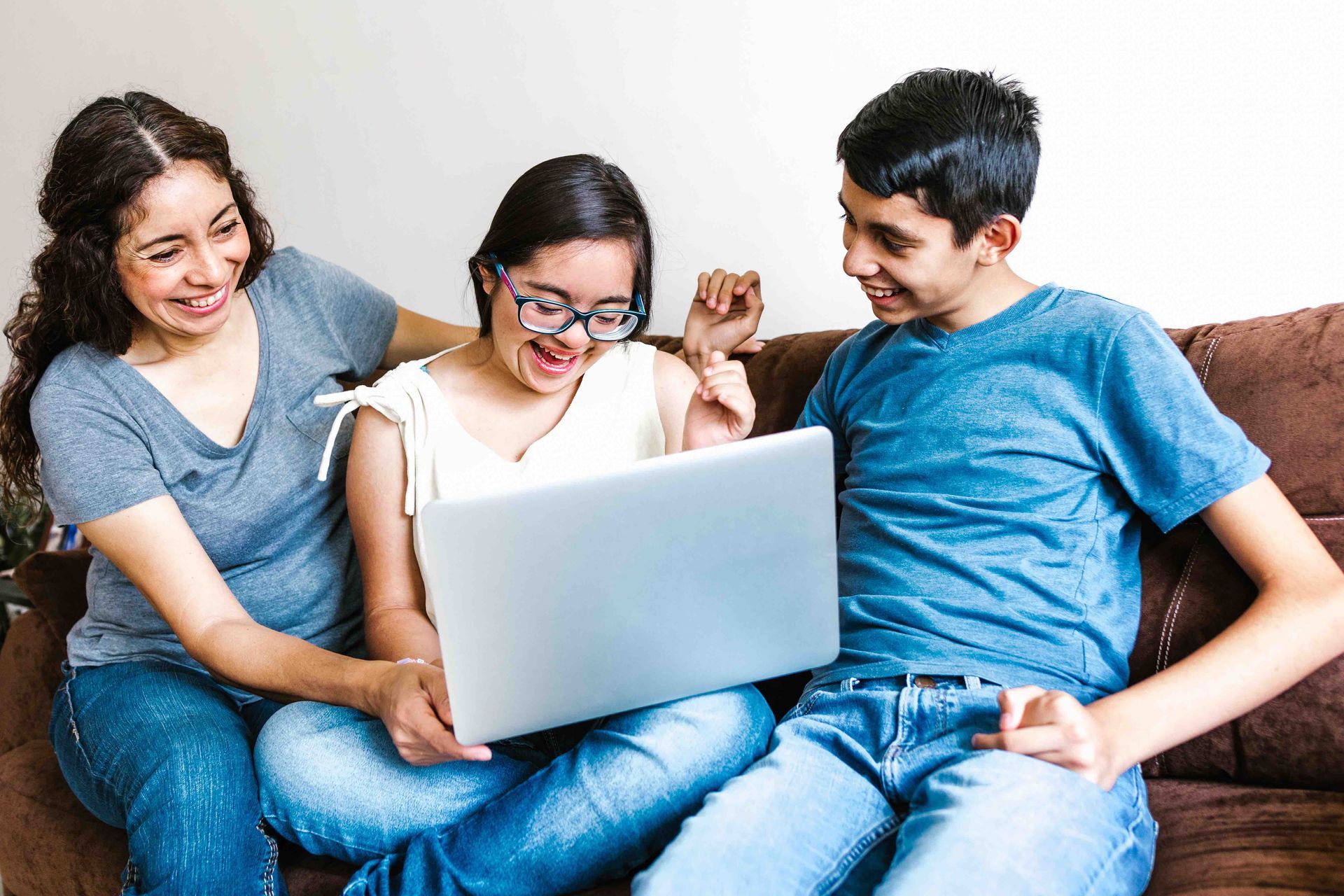Three people sitting together on a couch smile and laugh while looking at a laptop, showing a supportive home environment where individuals with intellectual and developmental disabilities engage in shared activities with caregivers.