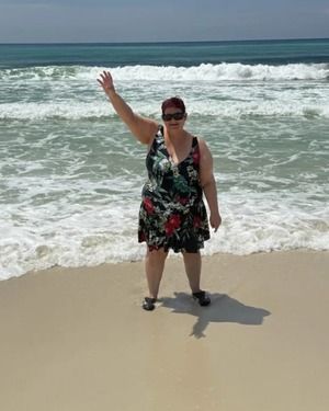 Woman on beach waves, wearing floral dress, waving her hand, sunny day.