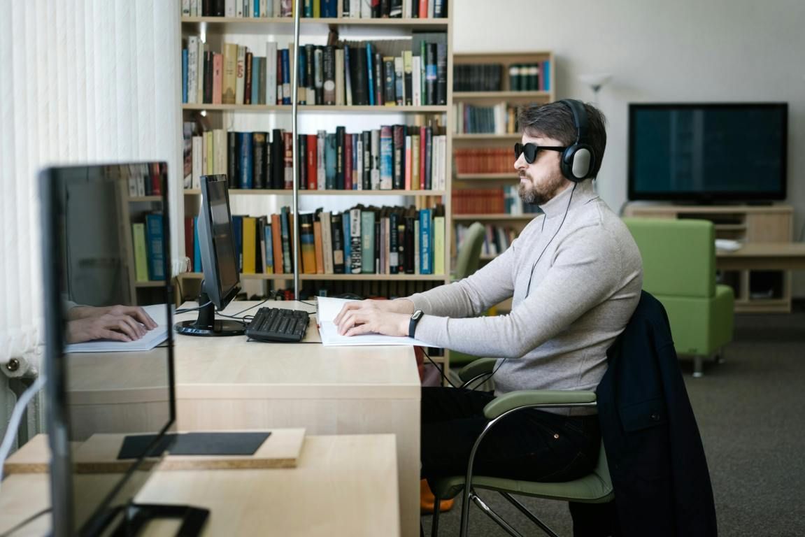 Man with headphones and dark glasses at a computer in a library. He types, with bookshelves in background.