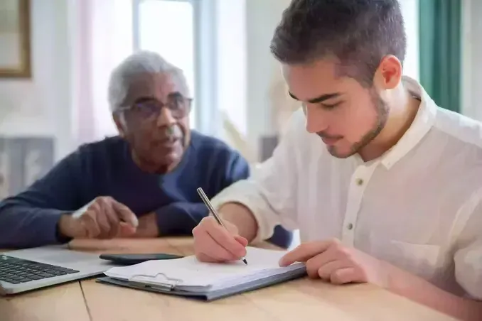 Young person writing on a clipboard, older person looking on; both at a table with a laptop.
