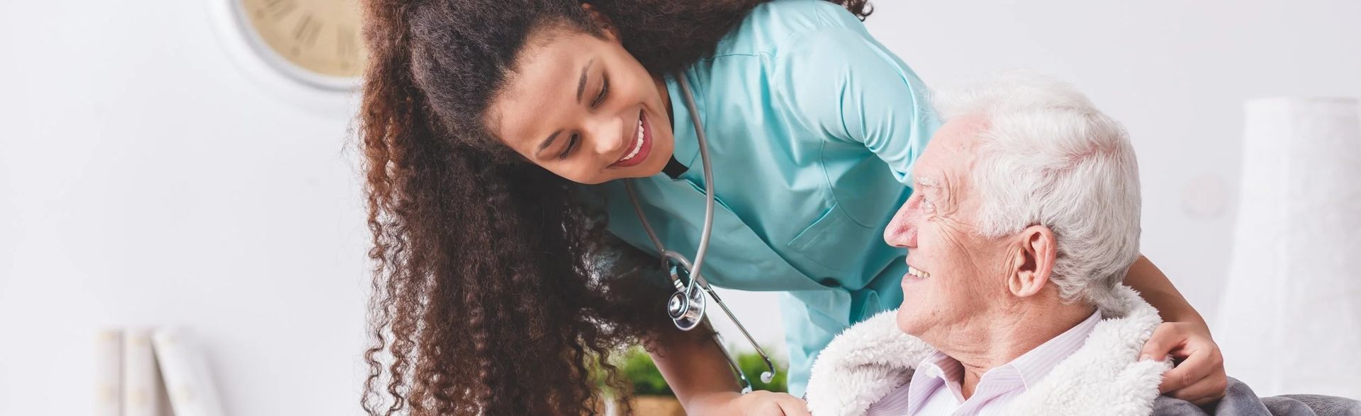 A healthcare worker smiles at an older adult. The worker wears a stethoscope and light blue scrubs.