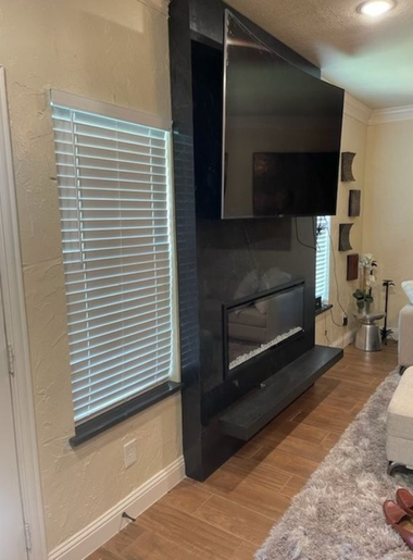 Living room with a large black fireplace, wall-mounted TV, window blinds, and hardwood floor