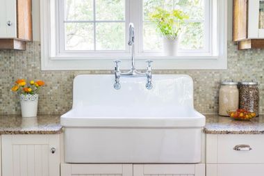 Bright kitchen sink under a window with flowers, granite countertops, and white cabinets