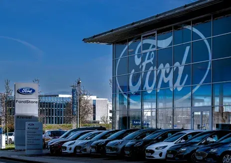 A row of cars parked outside a Ford dealership building featuring a large logo on its glass facade under a blue sky.