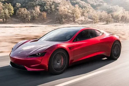 A red Tesla Roadster sports car driving fast on a paved road through a dry, hilly landscape.