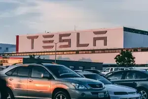 A Tesla factory building exterior with a large logo in the background, featuring parked cars in the foreground.