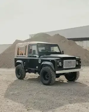 A black Land Rover Defender with a tan soft top parked on a gravel lot in front of large piles of earth.