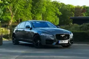 A dark gray Jaguar luxury sedan parked on a paved driveway surrounded by lush green trees and a small wooden bridge.