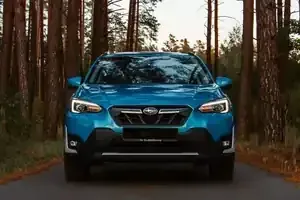 A bright blue Subaru Crosstrek parked on an asphalt path in a pine forest at sunset.