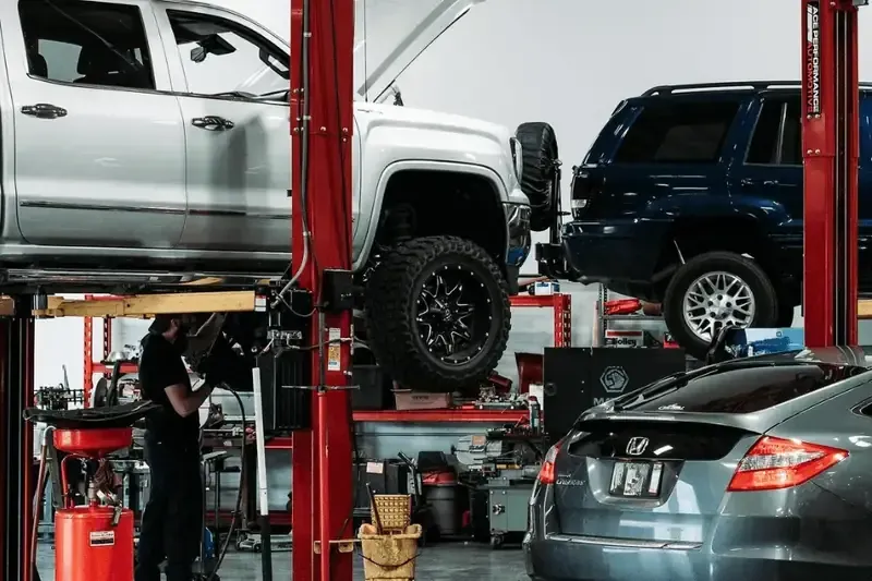 A worker repairs a white truck on a lift in a busy garage with a dark SUV and a grey sedan parked nearby.