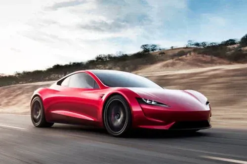A sleek red Tesla Roadster sports car driving along a desert road at high speed.