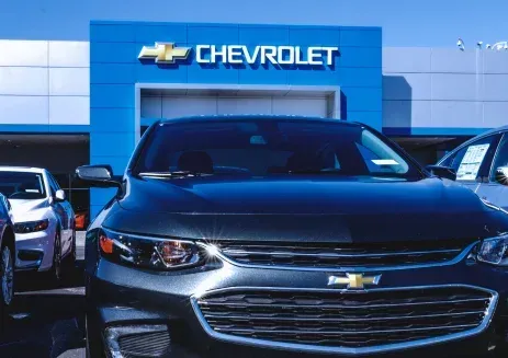 A dark grey Chevrolet sedan parked in front of a blue Chevrolet dealership building on a sunny day.