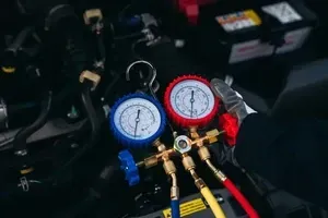 A mechanic's gloved hand adjusts an AC manifold gauge set connected to an automotive engine compartment.