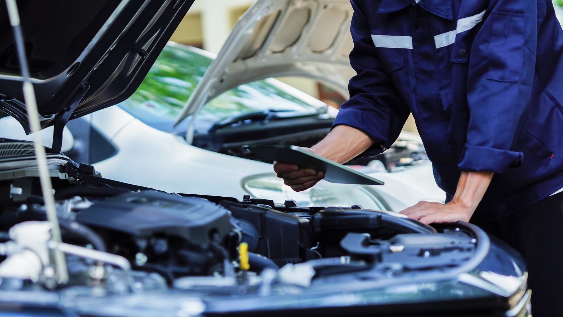 A mechanic in a blue uniform inspecting an open car engine while holding a tablet.
