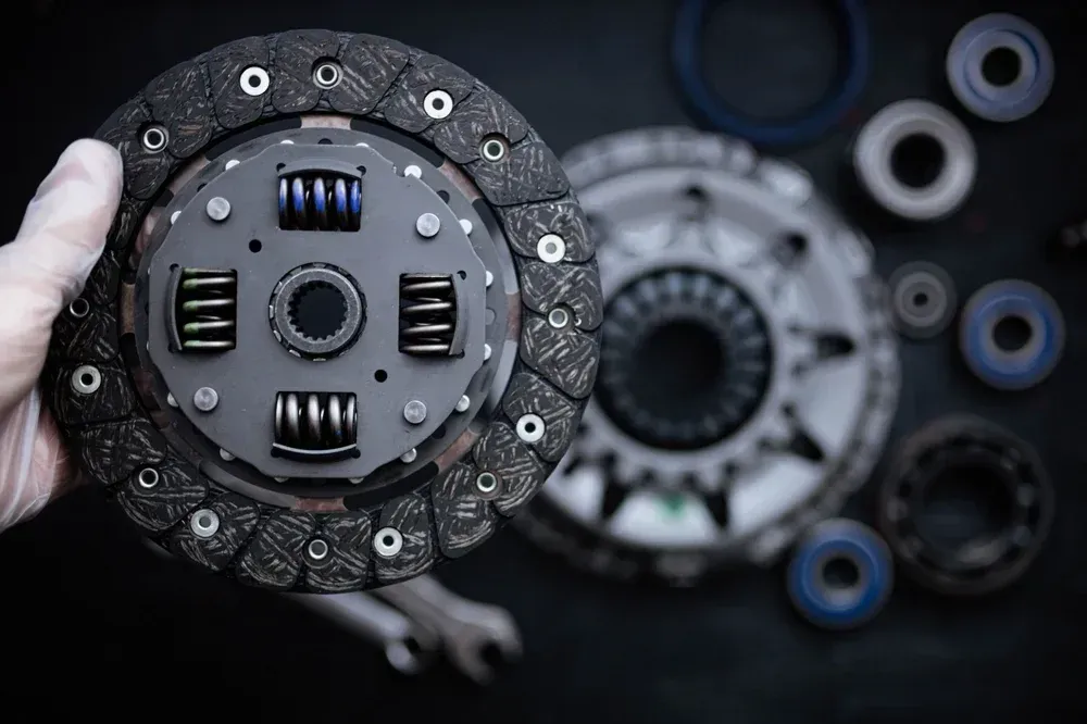 A gloved hand holds a circular automotive clutch disc with coiled springs, with other vehicle parts in the background.