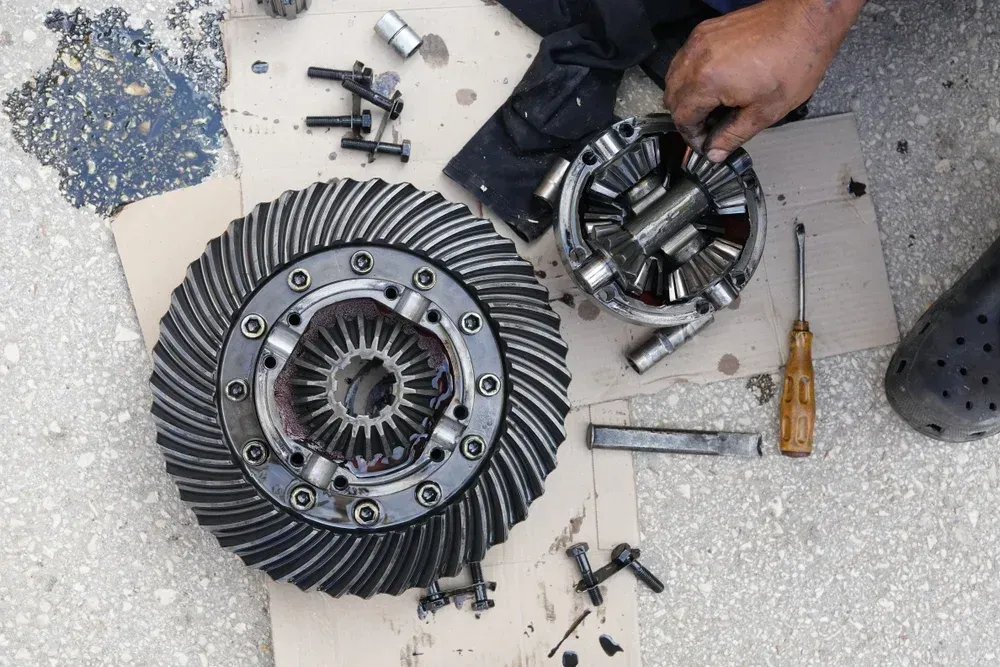 A mechanic works on a disassembled automotive differential assembly, with gears and tools laid out on cardboard.