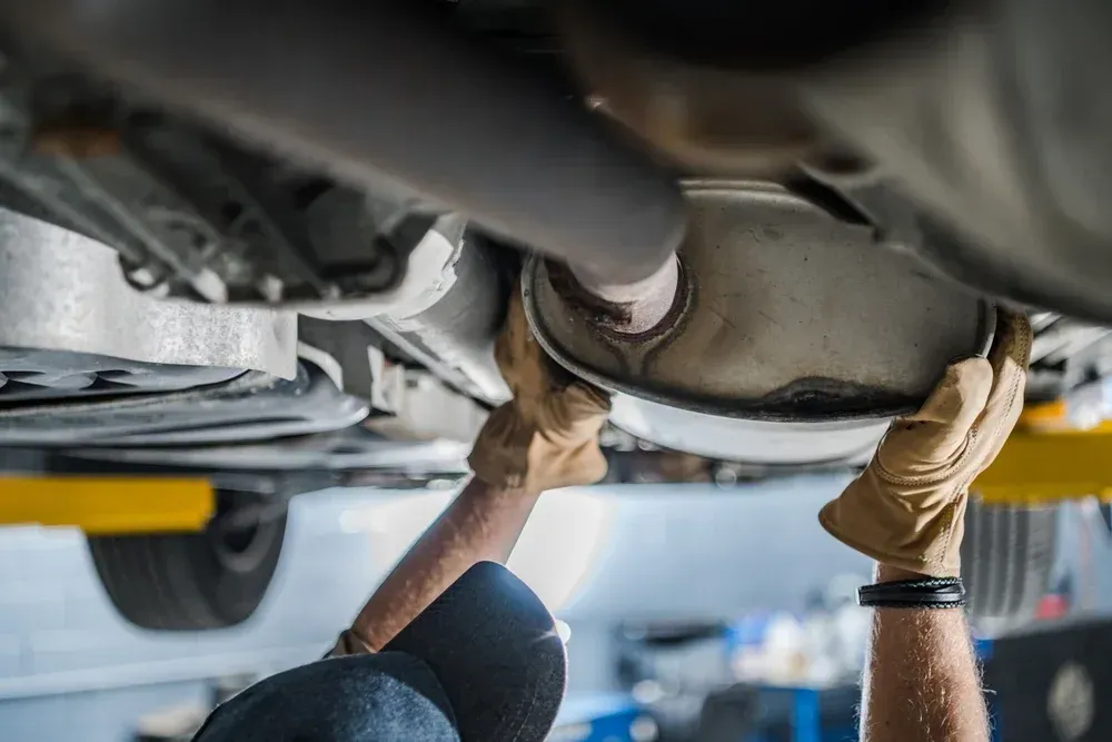 A technician in work gloves inspecting or repairing the muffler of a car lifted on a garage rack.