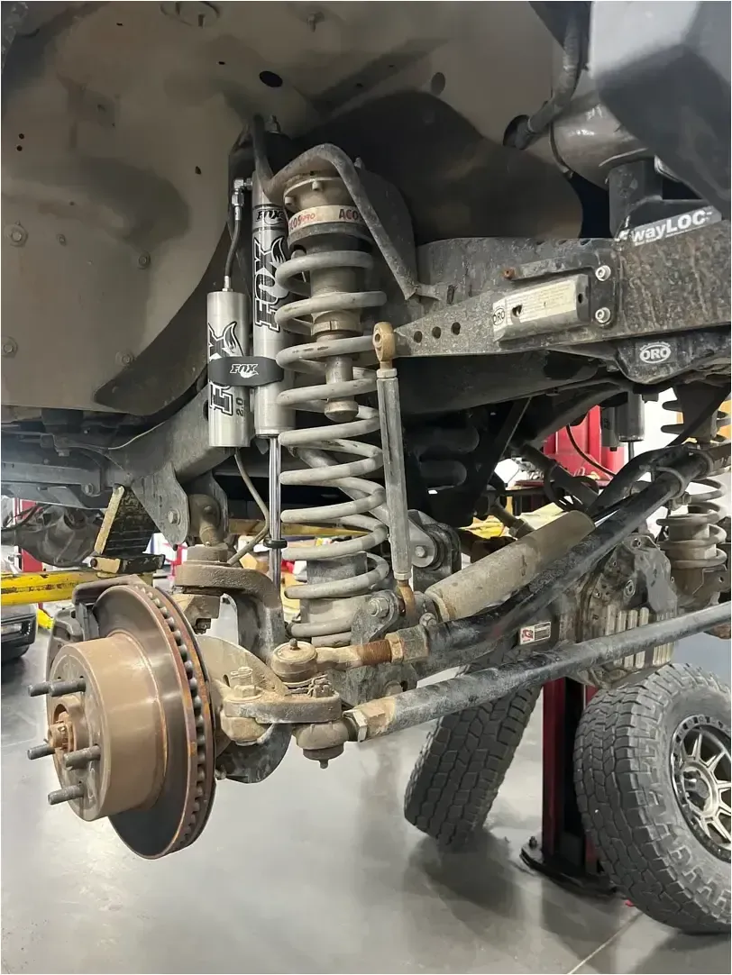 A mechanic in an auto repair shop works on a silver car lifted on a hydraulic hoist with its hood open.