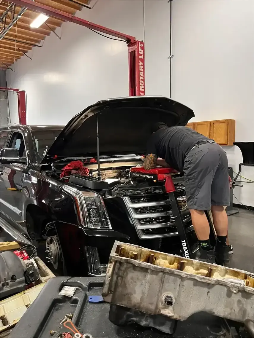 Two mechanics work together to lift an engine out of a black car using a hydraulic engine hoist in an auto shop.