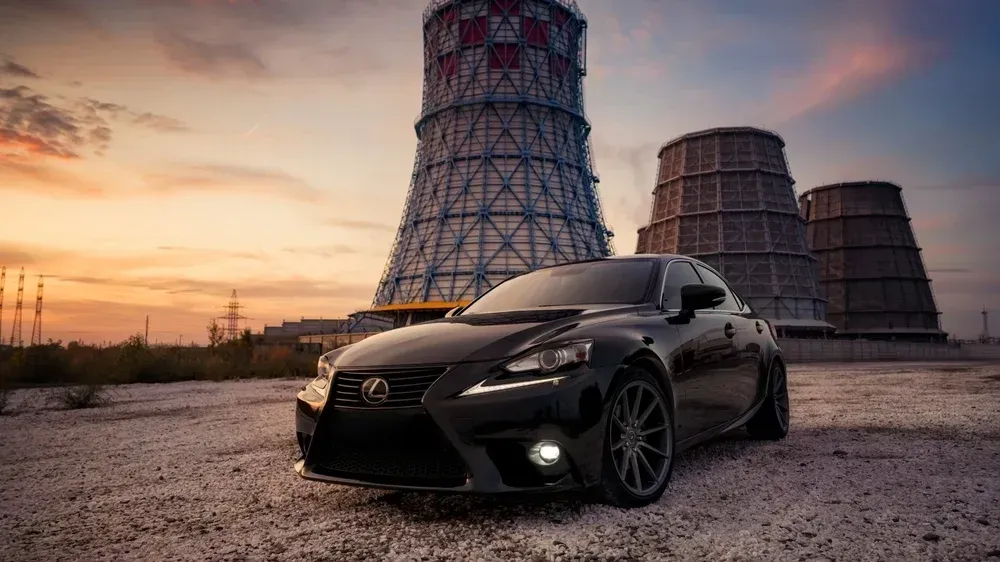 A black Lexus car parked on gravel in front of three large industrial cooling towers at sunset.
