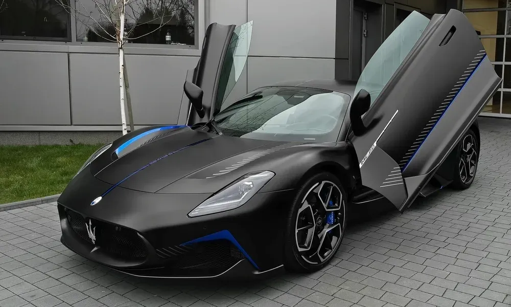 Black Maserati MC20 Cielo with butterfly doors open, parked on a paved driveway in front of a modern building.