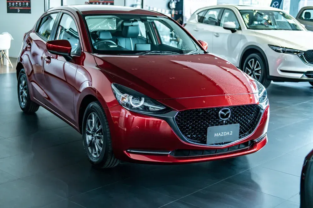 A red Mazda 2 sedan parked in a bright, modern car dealership showroom.