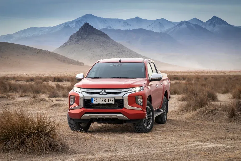 A red Mitsubishi pickup truck parked in a dry, sparse landscape with snow-capped mountains in the background.