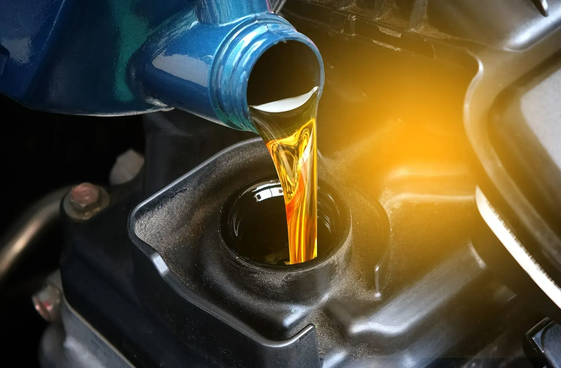 Golden motor oil being poured from a blue plastic container into a car engine.