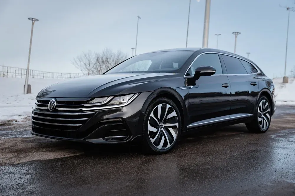 A dark grey Volkswagen Arteon station wagon parked on wet asphalt with a snow-covered bank in the background.