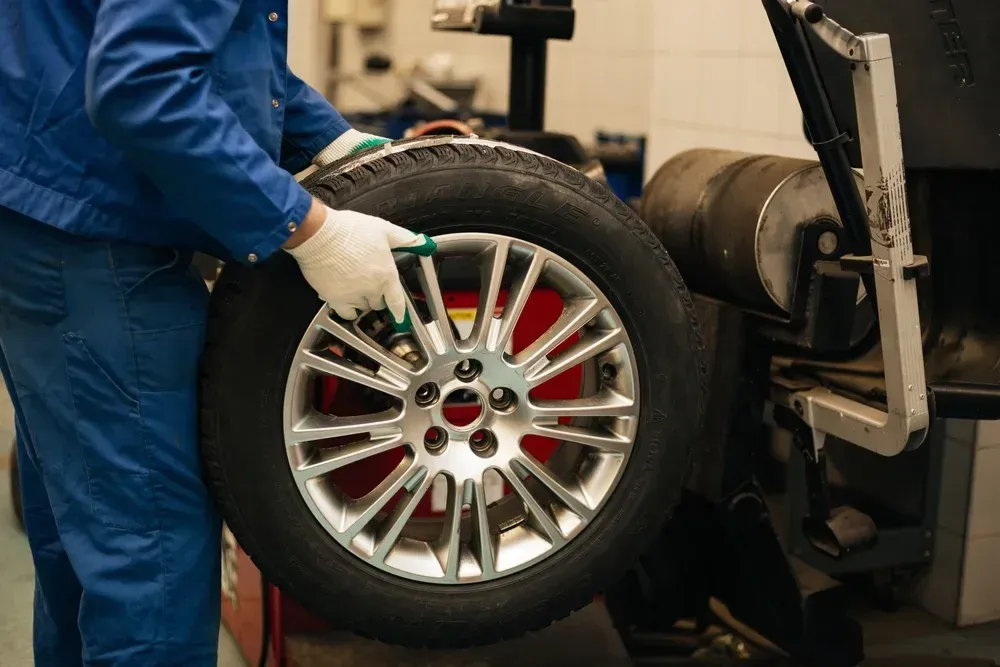 A technician in blue coveralls and gloves mounts a car tire onto a wheel balancing machine in an auto repair shop.