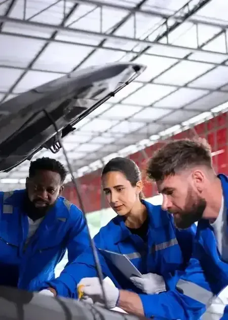 Three mechanics in blue uniforms examine the open hood of a car inside a brightly lit workshop.