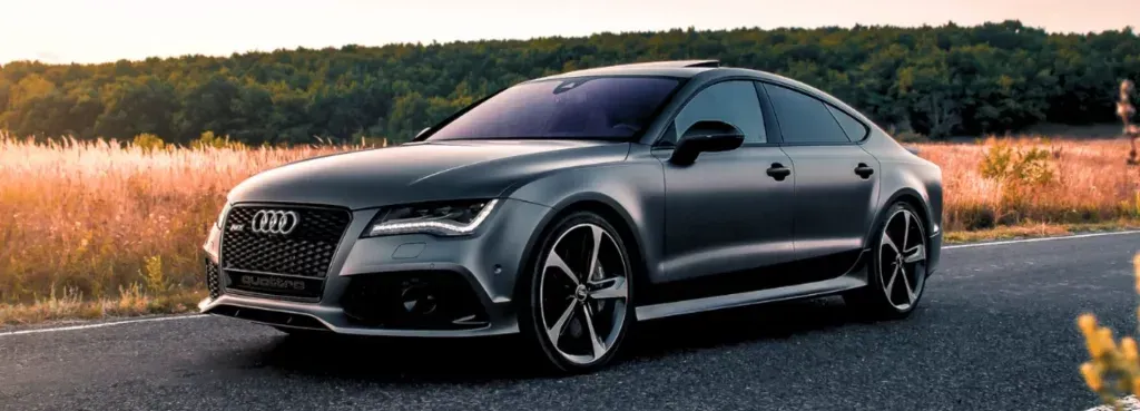 A matte grey Audi RS7 parked on a rural road at sunset, surrounded by tall grass.