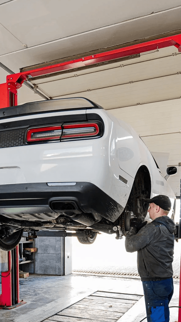 A mechanic works on the rear underside of a white Dodge Challenger lifted on a hydraulic hoist in an auto shop.