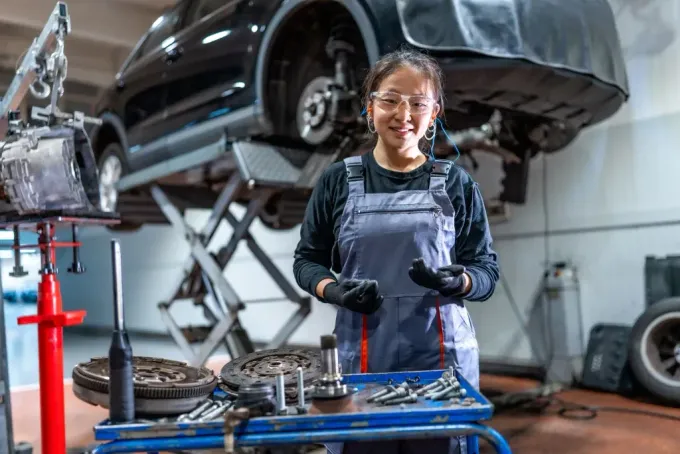 A mechanic wearing safety glasses and coveralls smiles while standing at a workbench in an auto repair shop with a car.