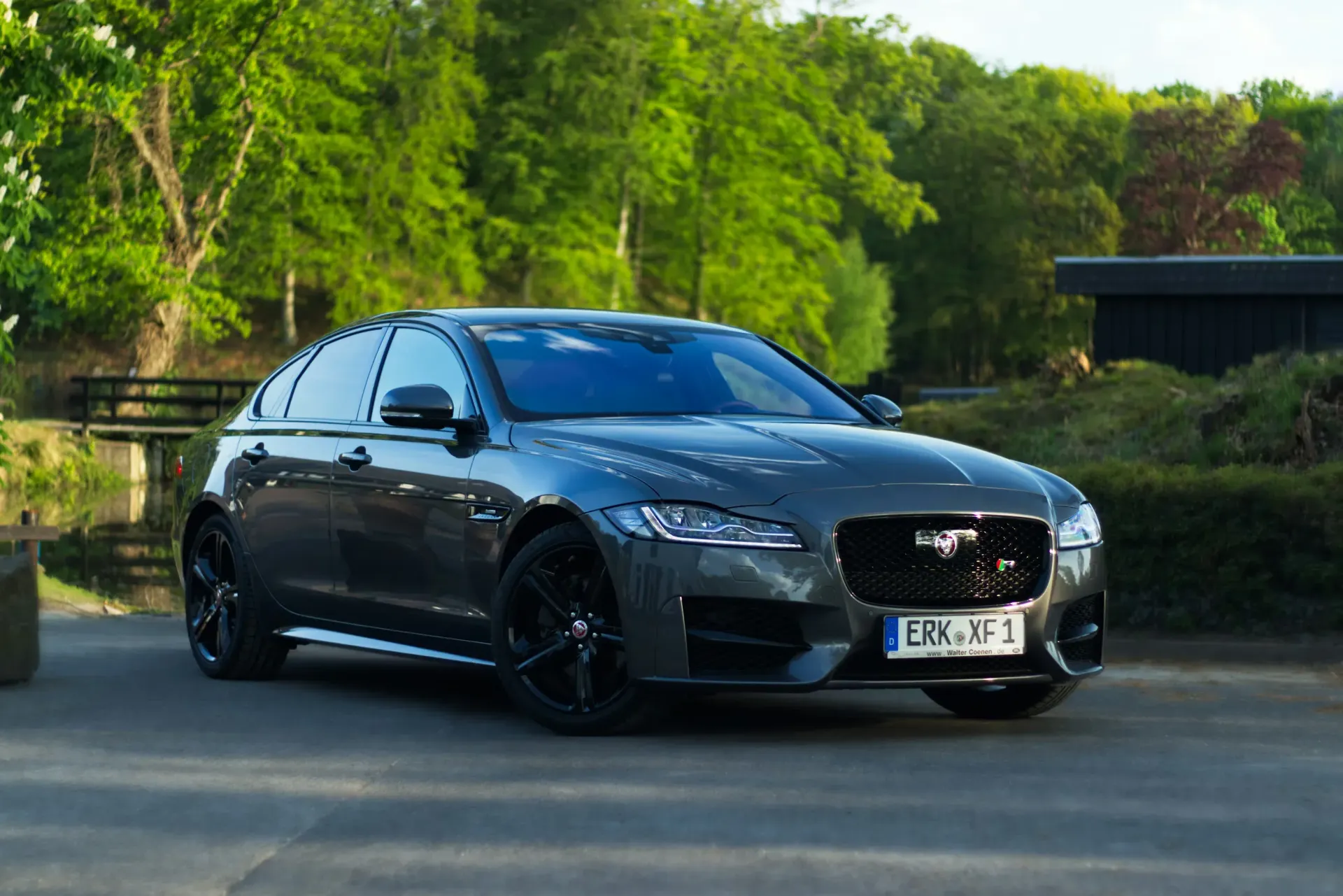 A dark gray Jaguar luxury sedan parked on a paved driveway surrounded by lush green trees and a small wooden bridge.
