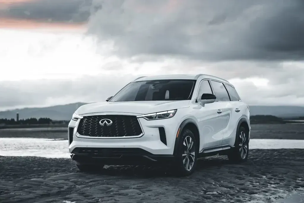 A white Infiniti SUV parked on a dark, rocky beach under a moody, overcast sky.