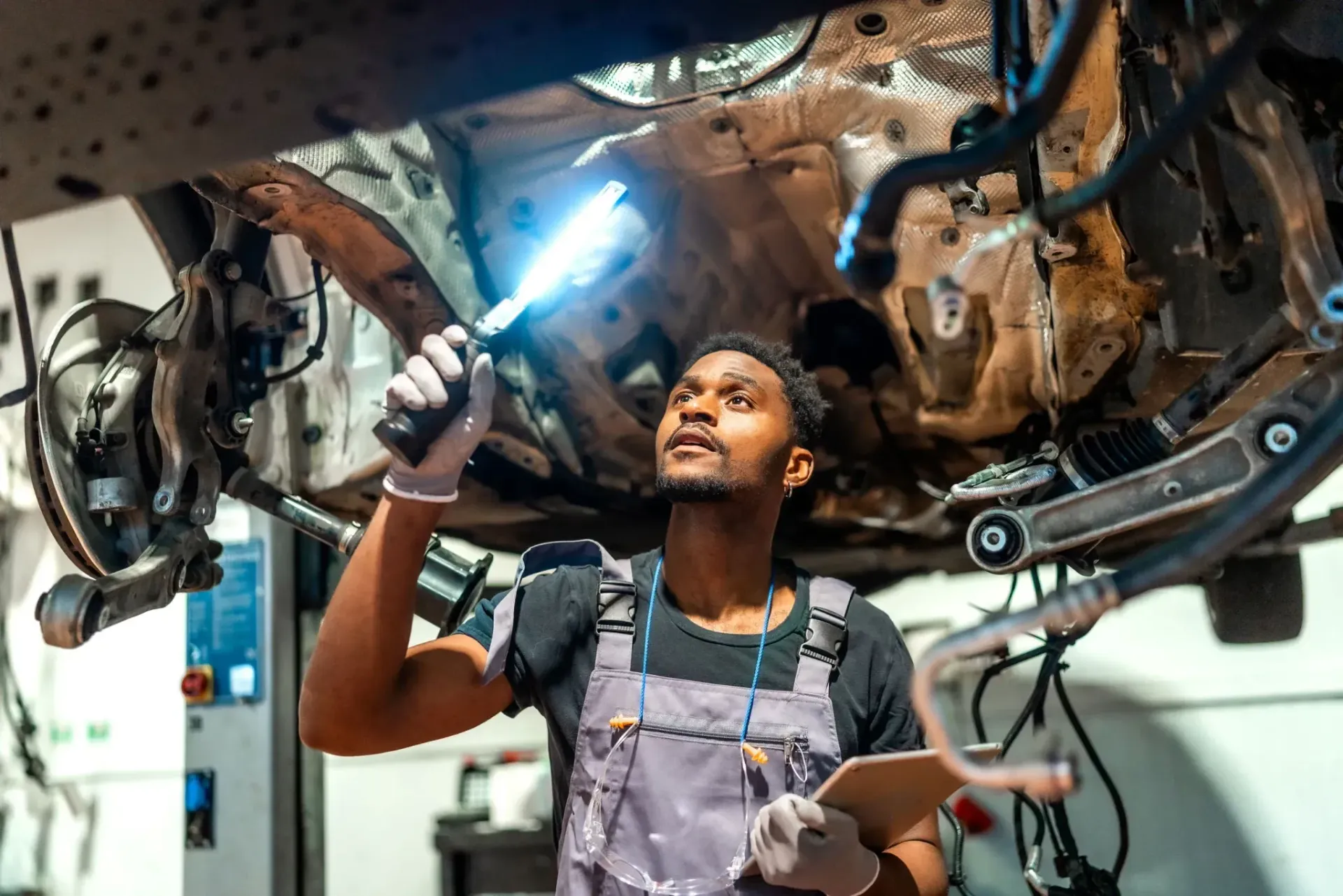 A mechanic wearing gray overalls holds a bright work light while inspecting the undercarriage of a vehicle in a garage.