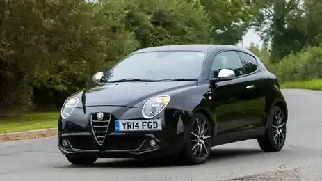 A black Alfa Romeo MiTo hatchback driving on a paved road surrounded by greenery.