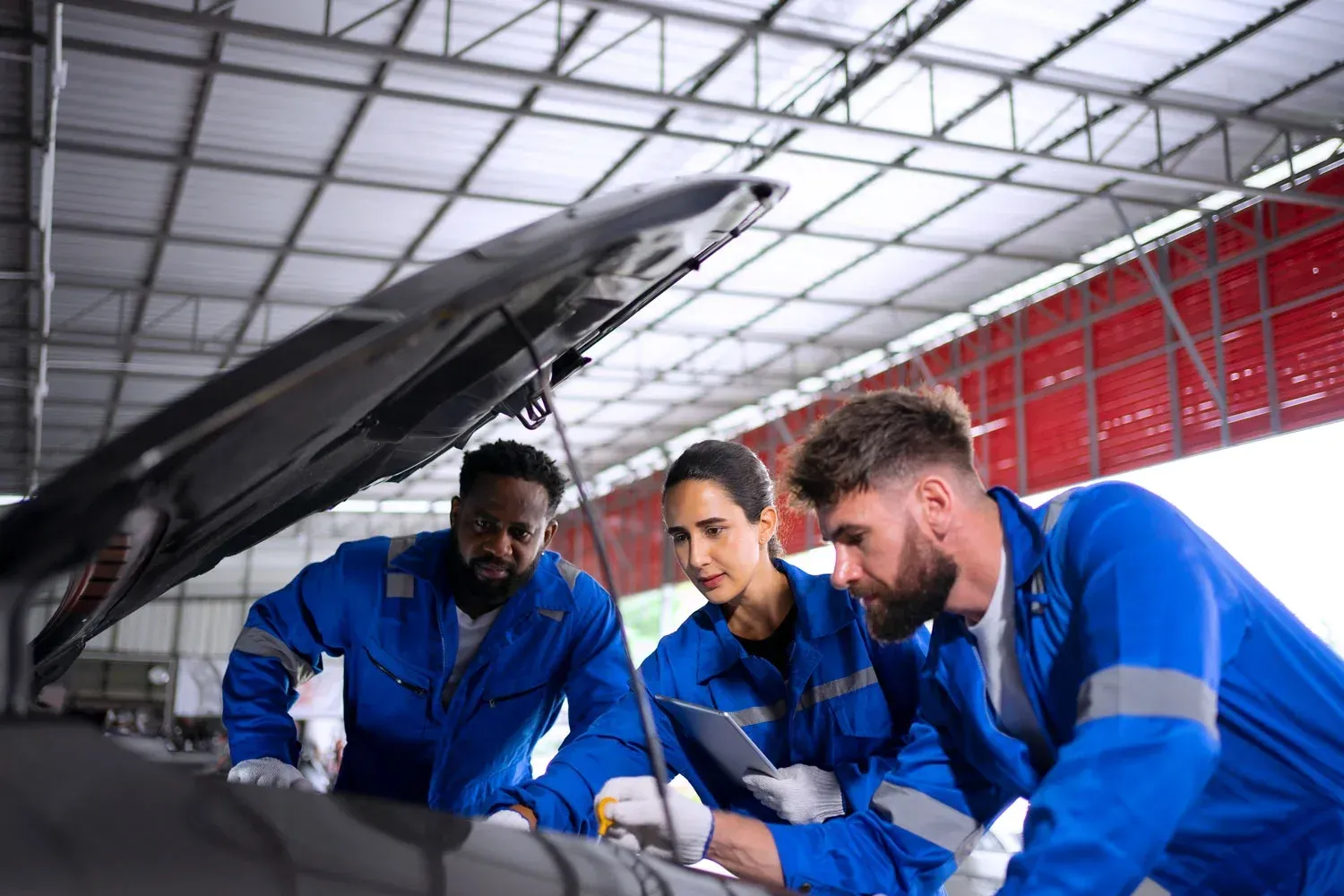 Three mechanics in blue uniforms inspect an open car engine in a professional garage setting, one holding a tablet.