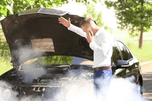 Man stands by a smoking car with an open hood, talking on a phone with one arm raised in frustration.
