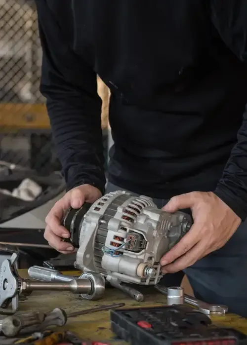 A person in a black long-sleeved shirt holds a metal car alternator in a workshop setting with tools on a table.