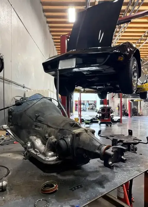 A removed car transmission sits on a metal workbench in a garage, with a classic sports car lifted on a hoist behind it.