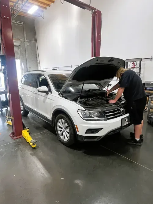 A technician works on the engine of a white SUV with its hood open in an auto repair shop.