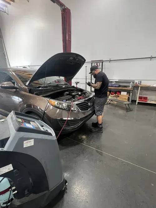 A mechanic works under the open hood of a brown SUV in a workshop, using diagnostic equipment connected to the engine.