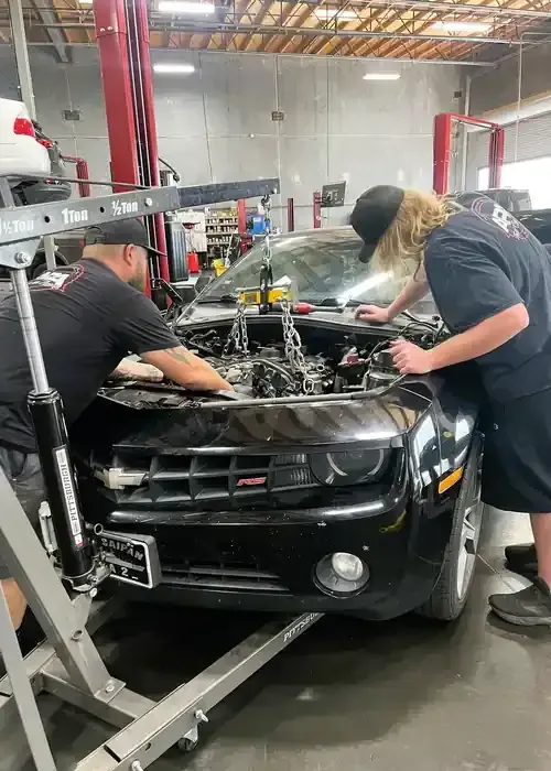 Two mechanics work together to lift an engine out of a black car using a hydraulic engine hoist in an auto shop.