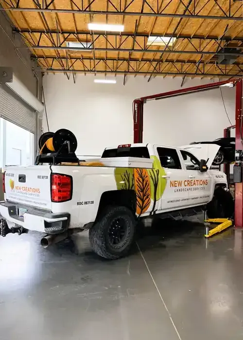 A white New Creations service truck with colorful branding parked in an auto repair shop under a car lift.